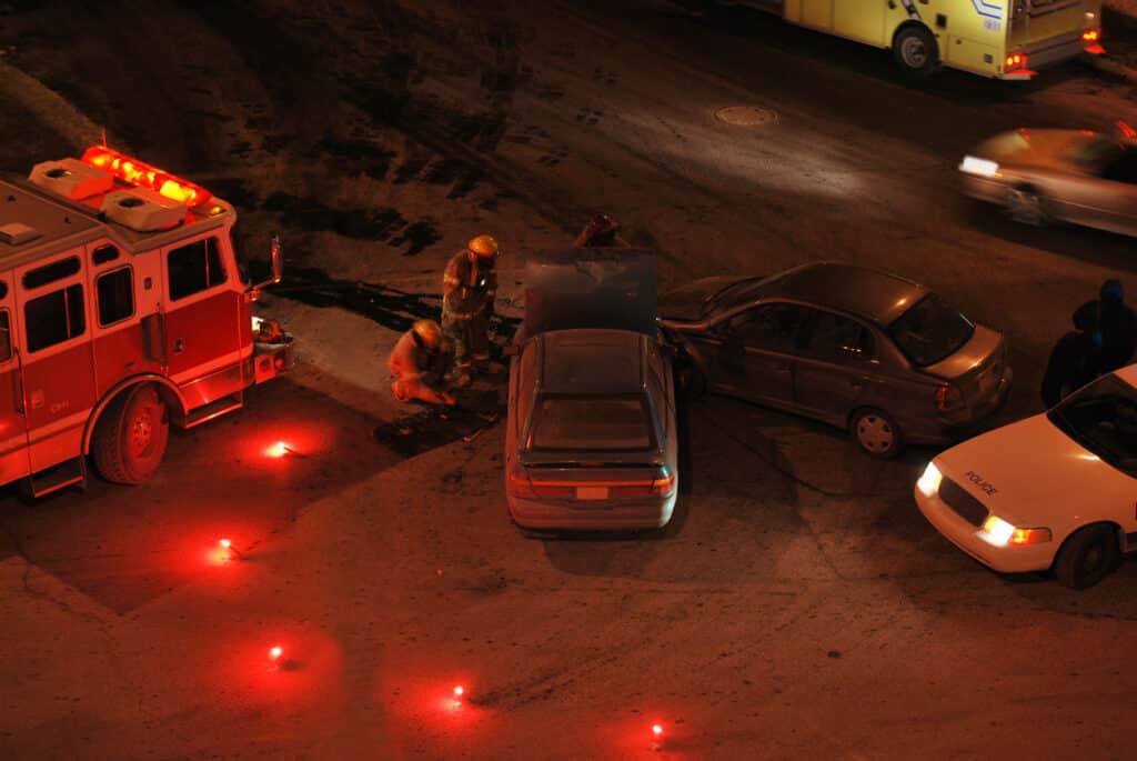Three cars in an accident at night time with a police car, firefighters, and ambulance partially visible.
