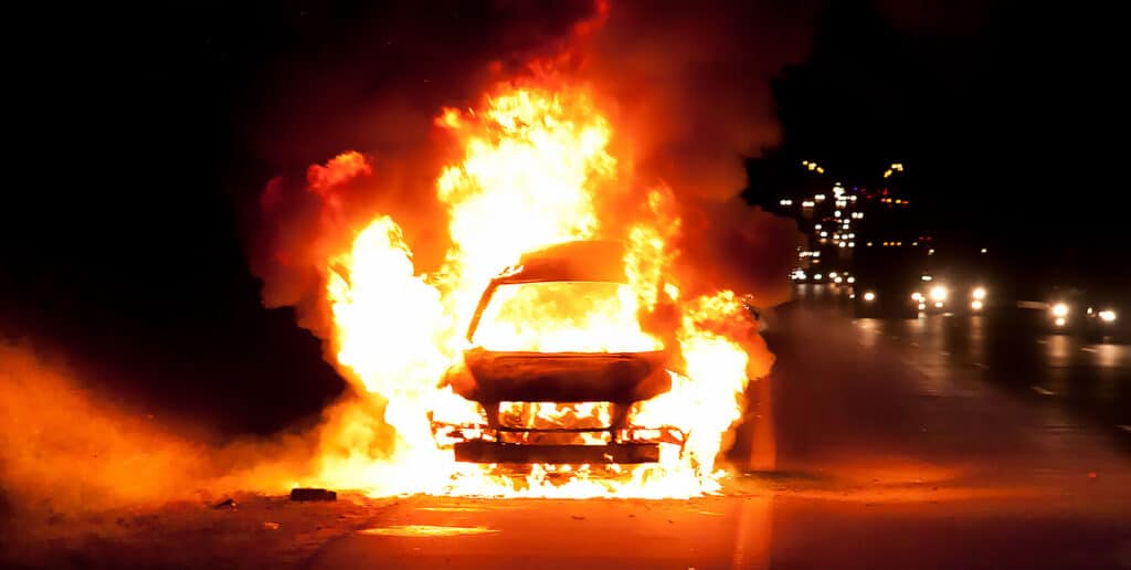 A car on fire on the shoulder of the highway at night.